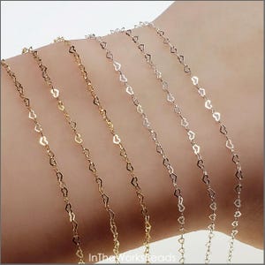 May include: Close-up of several delicate heart-shaped chain necklaces in gold and silver tones. The chains are draped across a person's arm, showcasing the intricate design and varying metallic finishes. The image is well-lit, highlighting the jewelry's details.