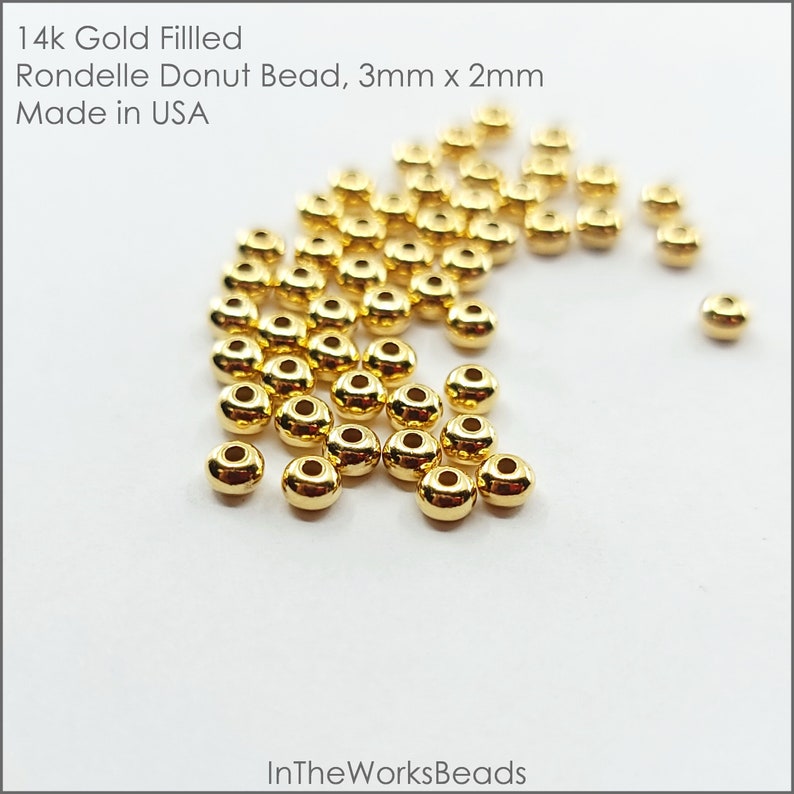 May include: A close-up of a pile of 14k gold filled donut beads, measuring 3mm x 2mm. The beads are shiny and have a smooth, round surface. Made in the USA.