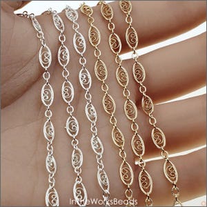 May include: Close-up of several delicate chains in silver and gold tones. The chains feature an oval link design with a decorative swirl pattern. The chains are held in a hand, showcasing the different colors and textures.