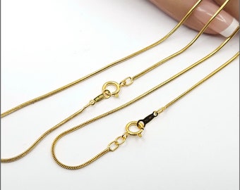 May include: Two gold snake chains with lobster clasps. The chains are laid out on a light pink surface.