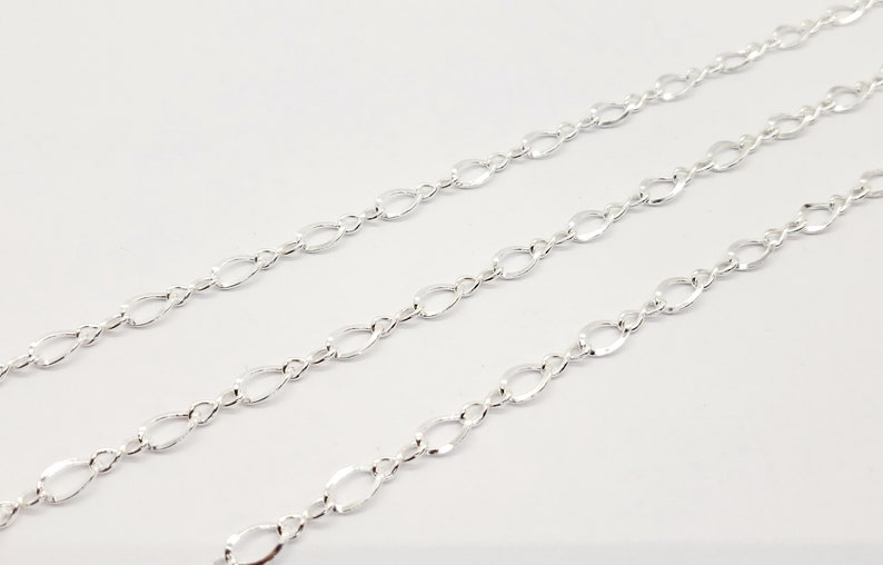 May include: Three strands of silver-coloured chain with an oval and figure-eight link design. The chains are arranged diagonally across a white background, creating a pattern of repeating shapes.