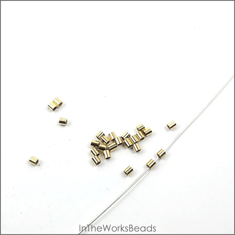 May include: A close-up of a pile of gold-colored crimp beads, some of which are strung on a white thread. The beads are small and cylindrical in shape.