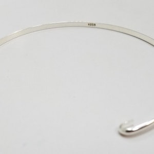Könnte beinhalten: Ein silbernes Armreif-Armband mit einem schlichten, offenen Design. Das Armband hat einen kleinen Hakenverschluss und ist innen mit "925" gekennzeichnet.