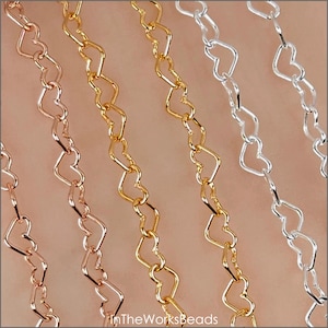 May include: Close-up of several heart-shaped chain necklaces in various metallic finishes: rose gold, gold, and silver. The chains are linked together in a repeating pattern. The image is well-lit, showcasing the details of the jewelry.
