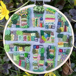 May include: A circular embroidery hoop with a gray felt background and colorful embroidery depicting a small town with houses, trees, and gardens. The embroidery is done in a variety of stitches and colors, creating a textured and detailed scene.