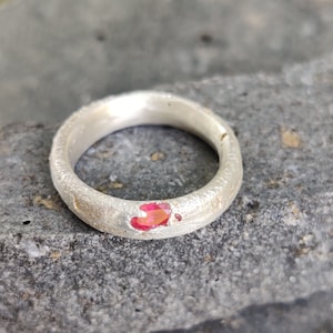 May include: A silver ring with a textured surface and a vibrant red gemstone. The ring is set against a gray background, showcasing the jewelry's design and color.