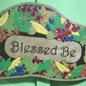 以下が含まれることがあります： 茶色の背景に白い筆記体で「Blessed Be」と書かれた木製の看板。看板は緑、赤、黄、青の色合いで描かれた葉、ベリー、ドングリで飾られています。