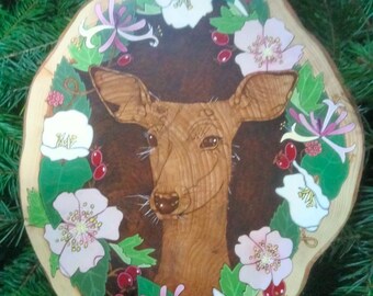 May include: A wooden slice with a painted deer head surrounded by pink and white flowers, green leaves, and red berries.