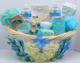 May include: A gift basket filled with self-care items. It includes blue and white bath products, a bracelet, a tumbler with the text "Not A Day Over Fabulous", and a box of Lindt Lindor white chocolate. The basket is decorated with blue and white flowers.