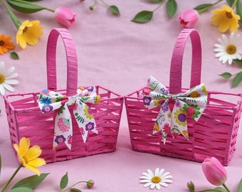 May include: Two bright pink woven baskets with matching handles and floral bows. The baskets are square-shaped and surrounded by pink and white flowers, including daisies, tulips, and other blooms, on a pink background.