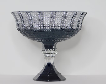 May include: An elegant, clear glass pedestal bowl with a dark blue base and rim. The bowl's rim is adorned with sparkling embellishments, and the dark blue section features intricate patterns. A decorative piece for serving or display.