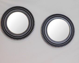 May include: Two round decorative mirrors. Each mirror has a black frame with silver accents and a reflective surface. The frames have a textured design with small, rounded silver elements. The mirrors are mounted on a white wall.