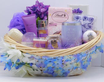 May include: A gift basket filled with purple and white items, including a tube of lotion, a perfume bottle, a jar of bath salts, a tumbler with text, and a box of chocolates. The basket is decorated with purple flowers and blue and white floral accents.