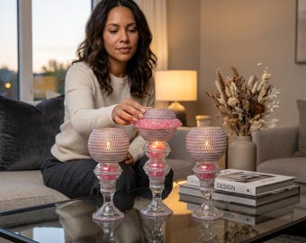 May include: Three decorative candle holders with pink accents. The candle holders have a clear glass base and a pink beaded top. The candles are lit, and the holders are on a glass table.