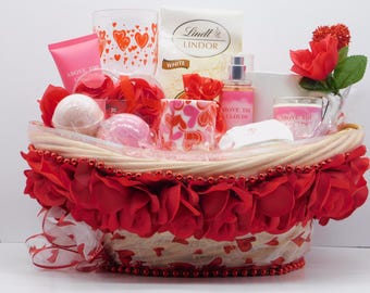 May include: A gift basket overflowing with Valentine's Day themed items. It includes a variety of products such as a pink lotion tube, a glass with heart designs, a bag of Lindt Lindor white chocolate, a bottle of perfume, a candle, and bath bombs. The basket is decorated with red roses and ribbon.