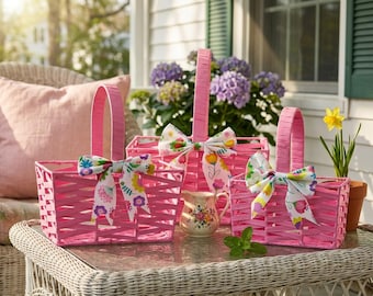 May include: Three pink woven baskets with handles and floral bows. A small floral pitcher and a potted daffodil are also present. The scene is set on a wicker table with a pink pillow and purple flowers in the background.