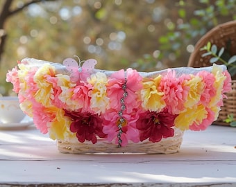 May include: A woven basket overflowing with pink, yellow, and burgundy artificial flowers. A pink butterfly decorates the top. The basket is on a white surface, with a blurred background of greenery.