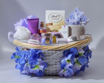 May include: A gift basket filled with self-care items. The basket contains a variety of products, including a purple lotion tube, a jar labeled "Love 100%", and a box of Lindor chocolates. The basket is decorated with blue flowers and a ribbon.