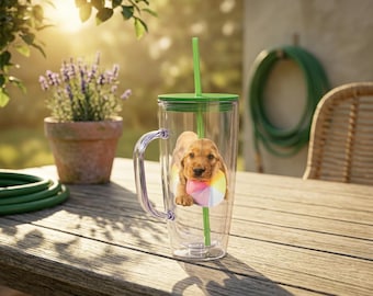May include: A clear tumbler with a green lid, handle, and straw. Inside the tumbler is a golden puppy wearing a colorful beach ball around its neck. The tumbler sits on a wooden table with a potted plant and a garden hose in the background.