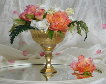 May include: A gold-colored, ornate pedestal bowl filled with a floral arrangement of pink, peach, and white flowers, with green fern accents. The bowl sits on a mirrored surface, with a single flower and scattered petals nearby.