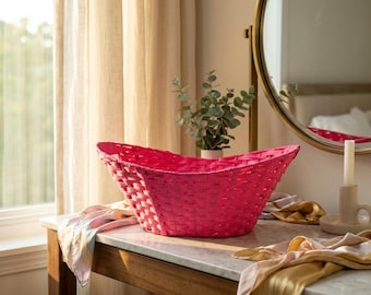 May include: A vibrant pink woven basket with a curved design sits on a marble-topped table. The basket is the focal point, with a textured weave. A gold-framed mirror and a candle holder are in the background.