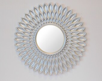 May include: A silver round mirror with a decorative frame made of overlapping petals. The mirror has a simple, modern design.
