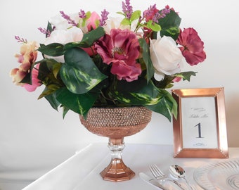 May include: A rose gold vase with a rhinestone trim holds a bouquet of pink, white, and purple flowers. A rose gold picture frame with the text "Table No. 1" is on a white tablecloth.