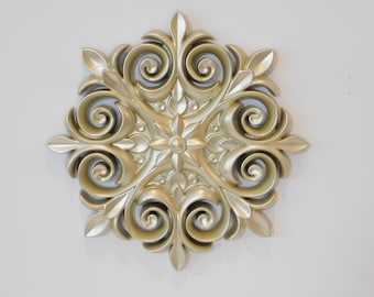 May include: A gold-colored decorative wall plaque with a floral design. The plaque has a raised, ornate pattern with swirls and leaves.