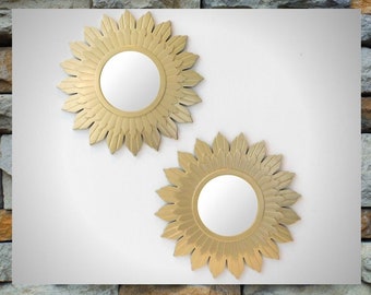May include: Two gold sunburst mirrors with a decorative, feathery design. The mirrors are round and have a reflective surface.