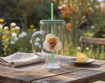 May include: A clear glass tumbler with a green lid, straw, and handle, featuring a Pomeranian puppy image. The tumbler sits on a wooden table with a lemon slice on a small plate and a linen napkin.