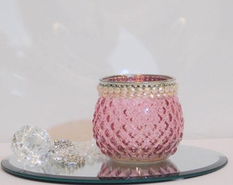 May include: Pink glass candle holder with a silver rim and a row of white beads around the top. The holder has a textured diamond pattern and is sitting on a silver mirrored surface.
