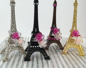 May include: Four miniature Eiffel Tower figurines with rhinestones and pink flower accents. The towers are silver, black, brown, and gold.