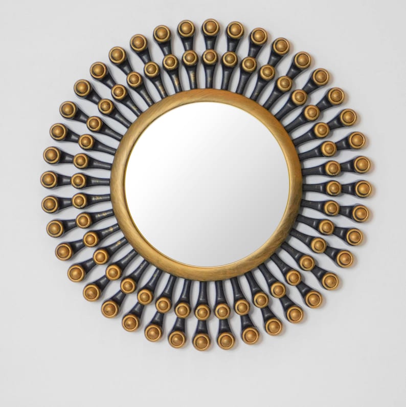 May include: A round decorative mirror with a gold-colored frame and a sunburst design. The sunburst features black and gold accents, creating a striking visual effect. The mirror is designed for wall mounting and adds a touch of elegance to any room.