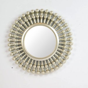 May include: A round, silver-colored mirror with a sunburst design. The mirror has a circular reflective surface surrounded by a frame of small, ball-shaped ornaments. The frame extends outward, creating a radial pattern.