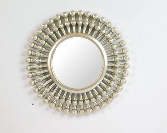 May include: A round, silver-colored mirror with a sunburst design. The mirror has a circular reflective surface surrounded by a frame of small, ball-shaped ornaments. The frame extends outward, creating a radial pattern.