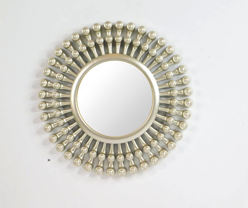 May include: A round, silver-colored mirror with a sunburst design. The mirror has a circular reflective surface surrounded by a frame of small, ball-shaped ornaments. The frame extends outward, creating a radial pattern.