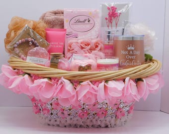 May include: A gift basket overflowing with pink items. It includes a tumbler with the text "Not A Day Over Fabulous", a candle, soap, lotion, chocolates, and a bracelet. The basket is decorated with pink flowers and rhinestones.
