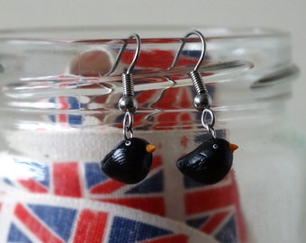 Può includere: Orecchini a forma di uccello nero con becco arancione e occhi bianchi. Sono appesi a ganci color argento. Lo sfondo include un barattolo di vetro e un nastro con un motivo rosso, bianco e blu.