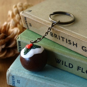 May include: A brown and white Christmas pudding charm with a green holly sprig on top, attached to a bronze key chain. The charm is sitting on top of a stack of three books with the titles "British Geology", "Wild Flowers", and "Weather".