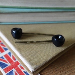 May include: Two black hair clips with gold bobby pins on a brown surface.