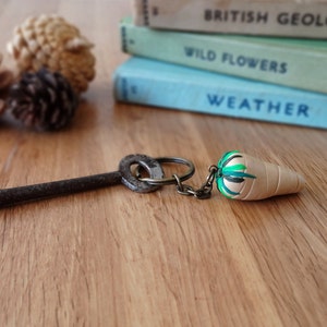 May include: A vintage key with a circular handle and a small carrot-shaped keychain. The keychain is beige with green and white accents. The key and keychain rest on a wooden surface, with books and pinecones in the background.