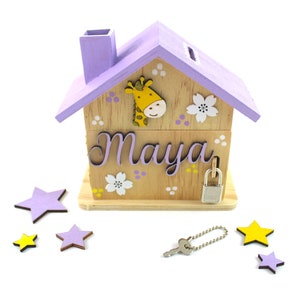 Könnte beinhalten: Ein hölzernes Sparschwein in Form eines Hauses mit einem violetten Dach, einem Giraffen-Design und dem Namen "Maya" auf der Vorderseite. Das Sparschwein hat ein Schloss und einen Schlüssel und ist von violetten und gelben Sternen umgeben.