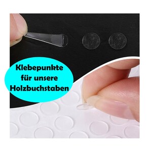 May include: A sheet of clear adhesive dots, with a hand holding one of the dots. The text "Klebepunkte f&uuml;r unsere Holzbuchstaben" is visible in the image.