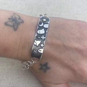 May include: A silver bracelet with a rectangular charm featuring various Halloween-themed symbols, including a ghost, crescent moon, star, and cross. The bracelet is on a person's wrist.