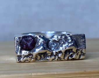 Può includere: Un anello d'argento con un design rettangolare strutturato. Una gemma viola scuro è incastonata nell'anello. L'anello ha un aspetto unico e organico, con una superficie irregolare e un'estetica moderna.