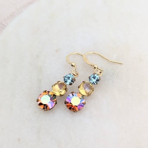 May include: Gold-coloured earrings with three crystals in a descending arrangement. The crystals display a gradient of colours, including orange, yellow, and blue. The earrings are set against a white marble surface.