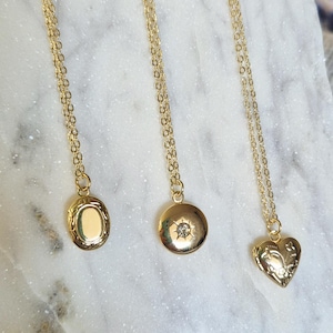 May include: Three gold necklaces with varying pendant designs: an oval locket, a round pendant with a central gem, and a heart-shaped locket. Each necklace features a fine chain. The pendants are presented on a white, marble-effect surface.