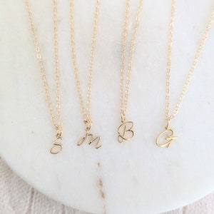 May include: Gold-coloured initial necklaces with delicate chains. The pendants feature the letters S, m, B, and G in a cursive script. The necklaces are displayed on a white marble surface.