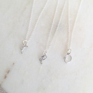 May include: Three silver necklaces with cursive initial pendants: T, P, and O. The pendants hang from fine silver chains. The necklaces are displayed on a white marble surface. The jewellery is simple and elegant.