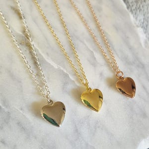 Puede incluir: Tres collares con colgantes en forma de coraz&oacute;n en plata, oro y oro rosa. Cada collar presenta una cadena delicada y un colgante de coraz&oacute;n liso y pulido. Los collares se exhiben sobre una superficie de m&aacute;rmol blanco y gris.
